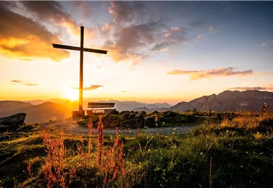 Ein wunderschöner Sonnenuntergang hinter einem Kreuz auf einem Berggipfel. Blumen und eine Bank im Vordergrund ergänzen die friedliche Landschaft.