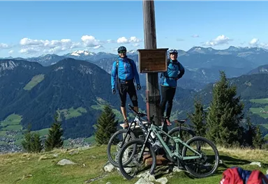 Two mountain bikers are standing at a summit cross with a view of the surrounding mountains. In the foreground, their bicycles are parked, while the landscape is green and hilly.