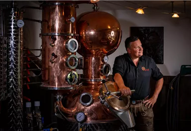 A man stands next to a large copper still in a distillery. The surroundings have a rustic atmosphere with various equipment and an industrial look.