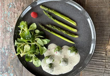 A black plate with green asparagus spears, fresh salad, and small vegetable dumplings in a creamy sauce. A raspberry decorates the dish.