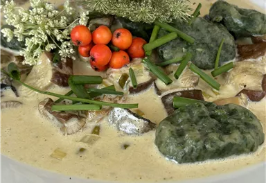 A dish with dark fish in a creamy sauce, garnished with green dumplings, fresh herbs, and red berries. The presentation is appealing and colorful.