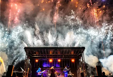 Eine Band spielt auf einer Bühne, während im Hintergrund ein spektakuläres Feuerwerk leuchtet. Die Szene ist lebhaft und festlich.