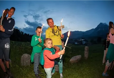 A group of children and adults is holding candles in a rural setting at dusk. In the background, mountains and a meadow are visible.