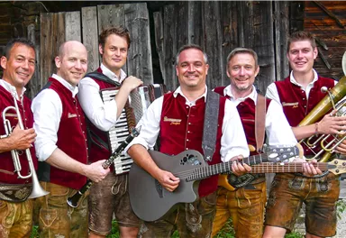 A group of six musicians in traditional Bavarian costume stands in front of a wooden building. They are holding various musical instruments, including an accordion, a guitar, and a trumpet.