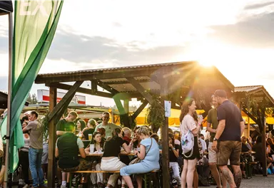 Ein belebter Markt mit vielen Menschen, die an Tischen sitzen und essen. Die Sonne scheint und sorgt für eine angenehme Atmosphäre.