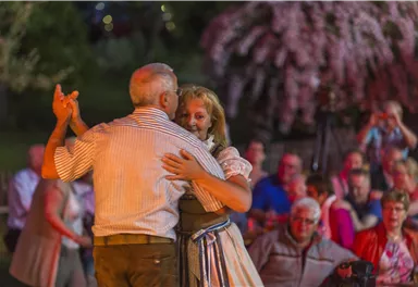 An older couple dances smiling at an event. In the background, spectators can be seen watching the performance.