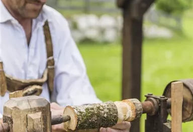 A craftsman is working on a lathe outdoors. In the background, green meadows and trees are visible.
