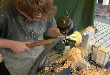 A craftsman works on a wood lathe and shapes a piece of wood. Sawdust flies through the air and covers the work surface.