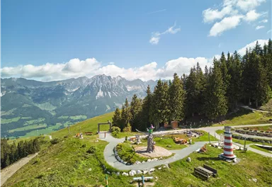 Eine wunderschöne Berglandschaft mit Wiesen und Bäumen. Im Vordergrund sind verschiedene Freizeitangebote und Skulpturen zu sehen.