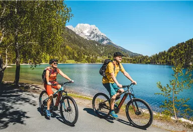 Zwei Radfahrer fahren an einem klaren blauen See entlang, umgeben von Bergen und Bäumen. Die Sonne scheint und der Himmel ist blau.