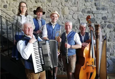 Six musicians in traditional attire stand in front of a stone wall. They hold various instruments such as accordion, clarinet, and harp.