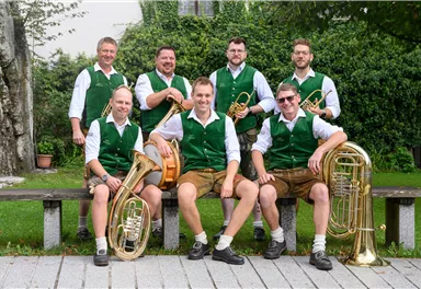 Eine Musikergruppe sitzt im Freien auf einer Bank. Sie tragen grüne Westen und spielen verschiedene Blasinstrumente.