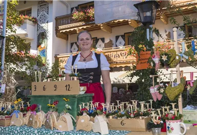 A woman in traditional attire stands at a market stall. The stall is decorated with colorful flowers and handcrafted items.