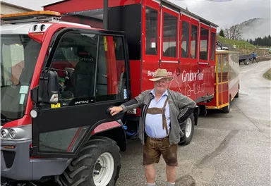 Ein Mann in traditioneller bayerischer Tracht steht neben einem Traktor mit einem roten Anhänger. Im Hintergrund sind sanfte Hügel und bewölkter Himmel zu sehen.