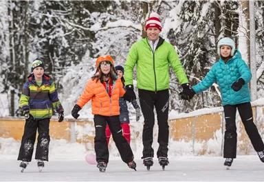 Eine fröhliche Gruppe von Kindern und einem Erwachsenen geht Schlittschuhlaufen auf einer eisbedeckten Fläche. Die Umgebung ist winterlich mit schneebedeckten Bäumen.