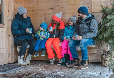 Eine Familie sitzt gemütlich in einer Holzhütte. Sie genießen heiße Getränke und tragen winterliche Kleidung.
