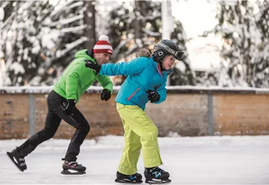 Zwei Kinder, die auf einer Eisbahn schlittschuhfahren. Es liegt Schnee im Hintergrund und sie tragen winterliche Kleidung.