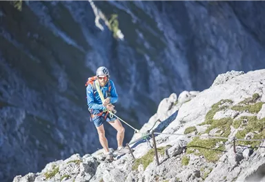 Ein Alpinist klettert an einer steilen Felswand. Er trägt Kletterausrüstung und fokussiert sich auf den Aufstieg.