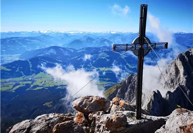 Ein beeindruckendes Bergpanorama mit einem Kreuz auf einem Gipfel. Im Hintergrund sind majestätische Berge und ein klarer Himmel zu sehen.