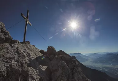 Ein Gipfel mit einem Kreuz und strahlendem Sonnenschein. Die umliegenden Berge sind klar und sichtbar im Hintergrund.