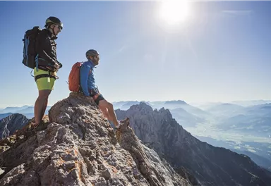 Zwei Bergsteiger sitzen auf einem Felsen und genießen die Aussicht auf die umliegenden Berge. Der Himmel ist klar und sonnig.