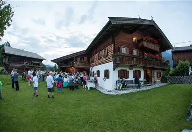A convivial gathering in front of a traditional wooden house. People are standing in the garden, enjoying the atmosphere at dusk.
