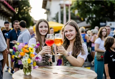 Zwei junge Frauen stoßen mit bunten Getränken an. Im Hintergrund sind Menschen und Marktstände zu sehen, die eine festliche Atmosphäre erzeugen.