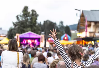 Ein lebhaftes Konzert im Freien mit einer Menge von Menschen. Ein Kind steht auf und hebt begeistert die Hände in die Luft.