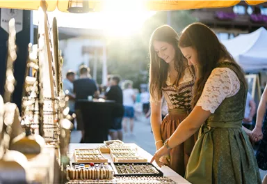 Zwei Frauen in traditioneller Kleidung schauen sich Schmuck auf einem Markt an. Im Hintergrund ist eine Sonnenstrahlung sichtbar, die eine gemütliche Atmosphäre schafft.