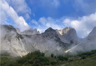 Ein beeindruckendes Bergpanorama mit felsigen Gipfeln und leichten Nebelschwaden. Der Himmel ist klar mit einigen Wolken.