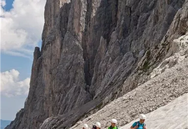 Eine Gruppe von Wanderern erklimmt einen felsigen Hang in den Bergen. Im Hintergrund sind hohe Felsformationen und ein klarer blauer Himmel zu sehen.