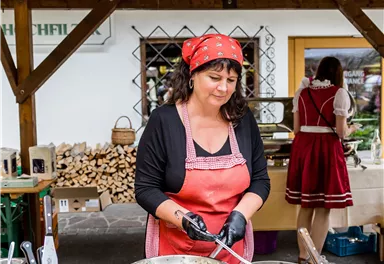 A woman in a cooking outfit is preparing food at a market stall. In the background, wooden stacks and other stalls are visible.