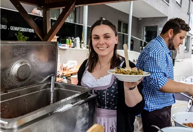 A woman in traditional attire is holding a plate of food in her hand. In the background, there is a man who is also preparing the food.
