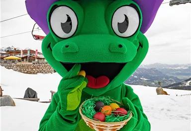 A cheerful mascot in a green suit with a purple hat stands in the snow holding a basket of colorful eggs. In the background, mountain landscapes and ski lifts are visible.