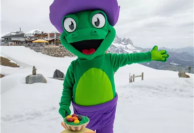A green mascot in a purple hat stands in the snow and smiles. It holds a bowl of food in one hand and points to the surroundings with the other hand.