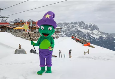 A green wizard in a colorful costume stands in the snow in front of a mountain landscape. In the background, a ski lift and a mountain cabin can be seen.