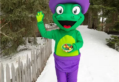 A colorful mascot stands in the snow and waves. It is wearing a purple hat and holds a bowl of colorful sweets.