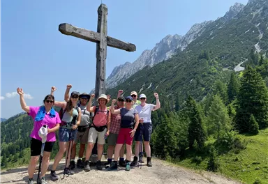 A group of hikers stands in front of a large wooden cross in the mountains. They are smiling and happily posing on a sunny day.