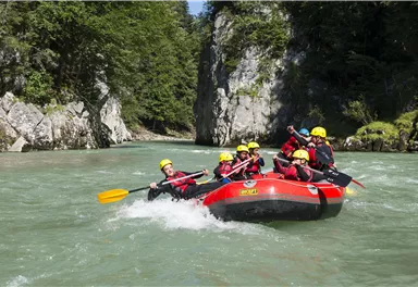Eine Gruppe von Menschen paddelt in einem roten Raft auf einem klaren Fluss. Umgeben von grünen Bäumen und Felsen bietet die Szenerie ein abenteuerliches Erlebnis in der Natur.