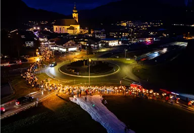 Eine festliche Veranstaltung bei Nacht mit vielen Menschen, die Lichter und Laternen tragen. Im Hintergrund ist eine Kirche und eine kleinräumige Stadtansicht zu sehen.