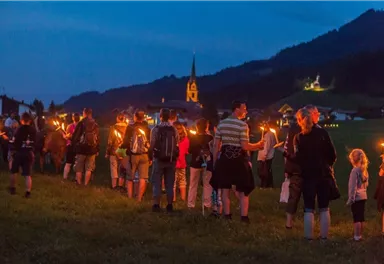 A group of people with torches walks through a meadow at dusk. In the background, a church and a picturesque landscape can be seen.