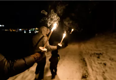 A group of people walks through the snow at night with torches. The surroundings are illuminated by the lights of the city in the distance.