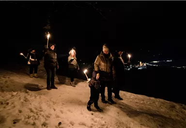 A group of people with torches is walking at night along a snow-covered path. In the background, the lights of a city can be seen.