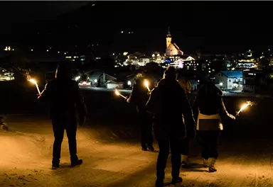 A group of people stands in the snow at night, holding torches in their hands. In the background, a lit city with a church can be seen.