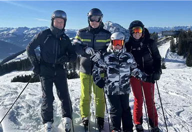 Eine Gruppe von vier Skifahrern steht auf einem schneebedeckten Hang. Im Hintergrund sind Berge und ein klarer Himmel zu sehen.