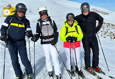 Eine Gruppe von vier Skifahrern steht auf einer verschneiten Piste. Der Himmel ist klar und blau.
