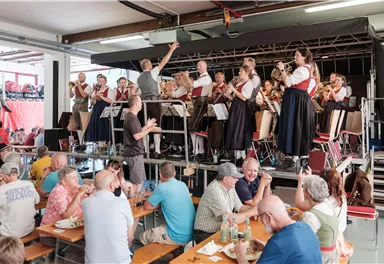 A brass band is playing on a stage while the audience sits at tables. The people are enjoying the food and the music in a sociable atmosphere.