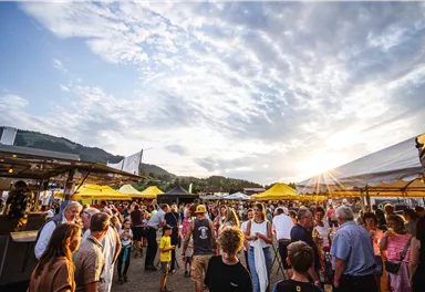 Ein lebendiger Markt mit vielen Menschen und bunten Ständen. Die Sonne scheint am Himmel und schafft eine fröhliche Atmosphäre.