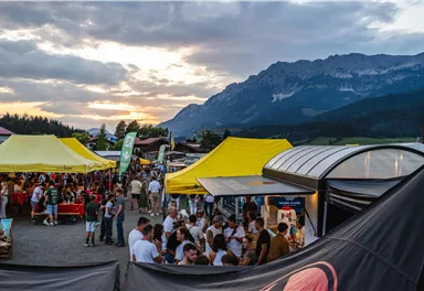 Eine belebte Veranstaltung mit vielen Menschen und bunten Zelten. Im Hintergrund sind imposante Berge und ein schöner Sonnenuntergang zu sehen.