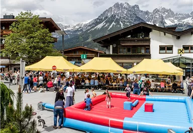 A lively outdoor event with yellow tents and many visitors. In the foreground, there is an inflatable play arena and a picturesque mountain backdrop.